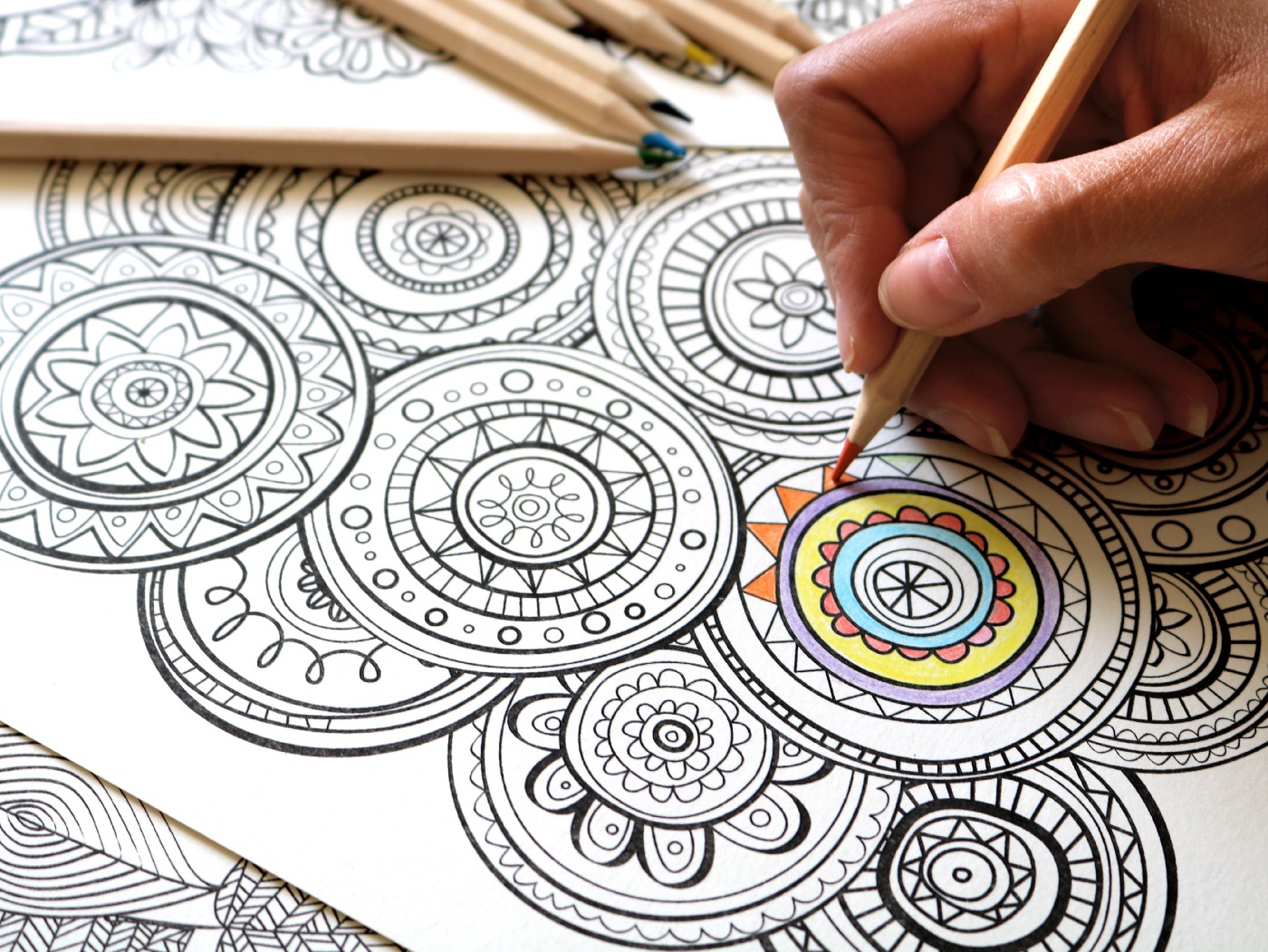 a hand coloring a detailed coloring page of a mandala with pastel colors as various colored pencils lay above