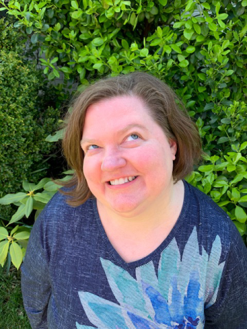 Image of woman wearing blue top standing in front of a green tree or bush