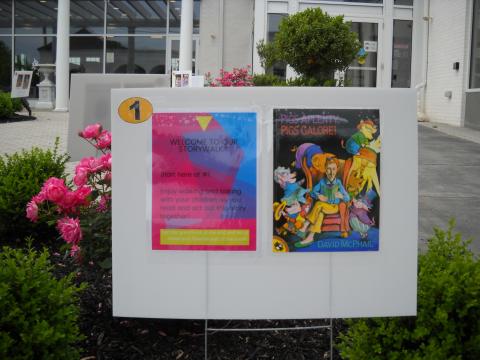 First sign of StoryWalk includes welcome page and cover of book. Rose bush in background.