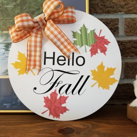 Circular sign with yellow-orange checkered bow, the wordsl Hello Fall, and yellow, red, and green leaves